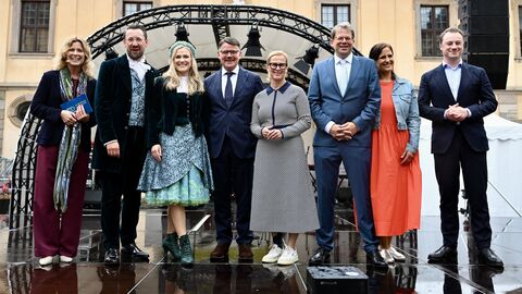 Eine Gruppe von acht festlich gekleideten Personen steht lächelnd nebeneinander auf einer nassen Open-Air-Bühne. Unter ihnen befindet sich eine Frau in einem auffälligen, blau-grünen Trachtenkleid mit Blumenkranz. Im Hintergrund ist ein historisches Schlossgebäude zu sehen.